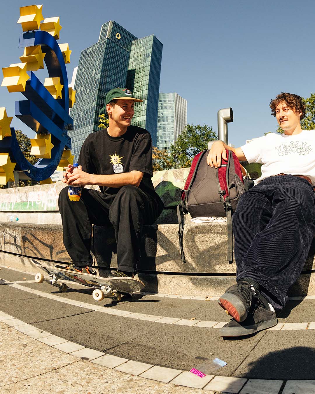 Two young skateboarders sitting in the front of skyscrapers, having a chat and laughing in the sun.