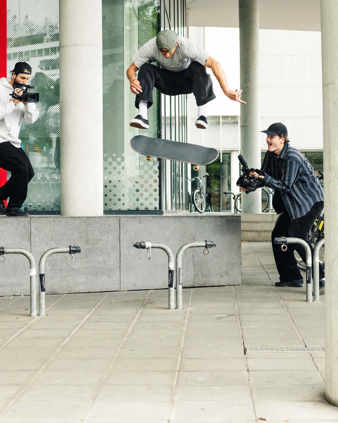 Still photo of skateboarder Marcel Weber doing a Frontside Kickflip down a gap while two videographers are capturing the trick.
