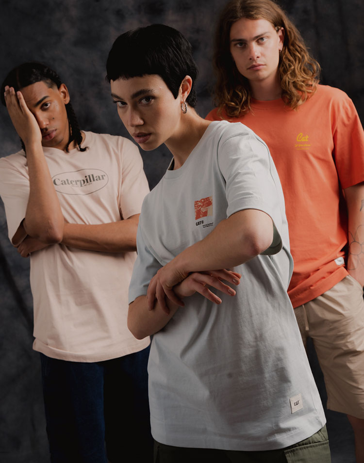 Studio photo of two young men with long hair in the background and a young woman with short hair in the center, all wearing Caterpillar t-shirts in pastel colors.
