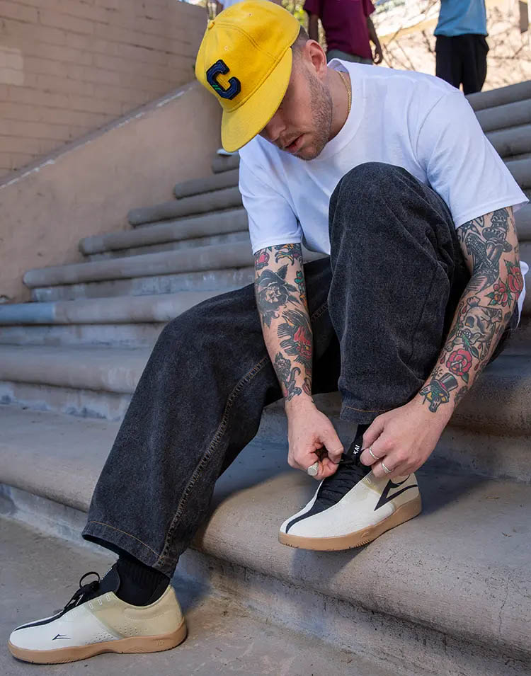 Picture of skateboarder Griffin Gass, sitting on a stairset with a yellow cap, white t-shirt and tattooed arms, lacing up his Lakai shoes