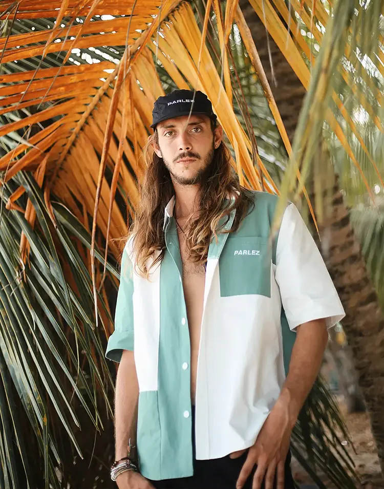 Handsome young man with long hair and beard, standing in front of yellowish palm tree leafs, looking into the camera and dressed with an non buttoned shirt and a black Parlez cap