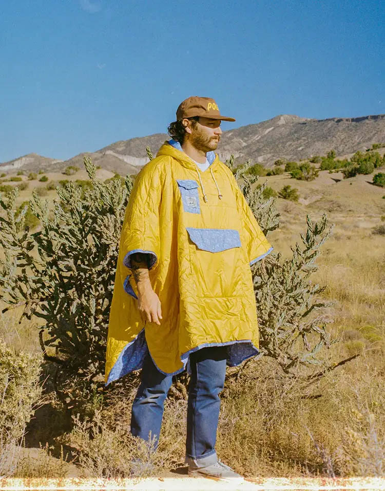 Male model with a cap and a yellow outdoor poncho in front of a blue sky desert scenery