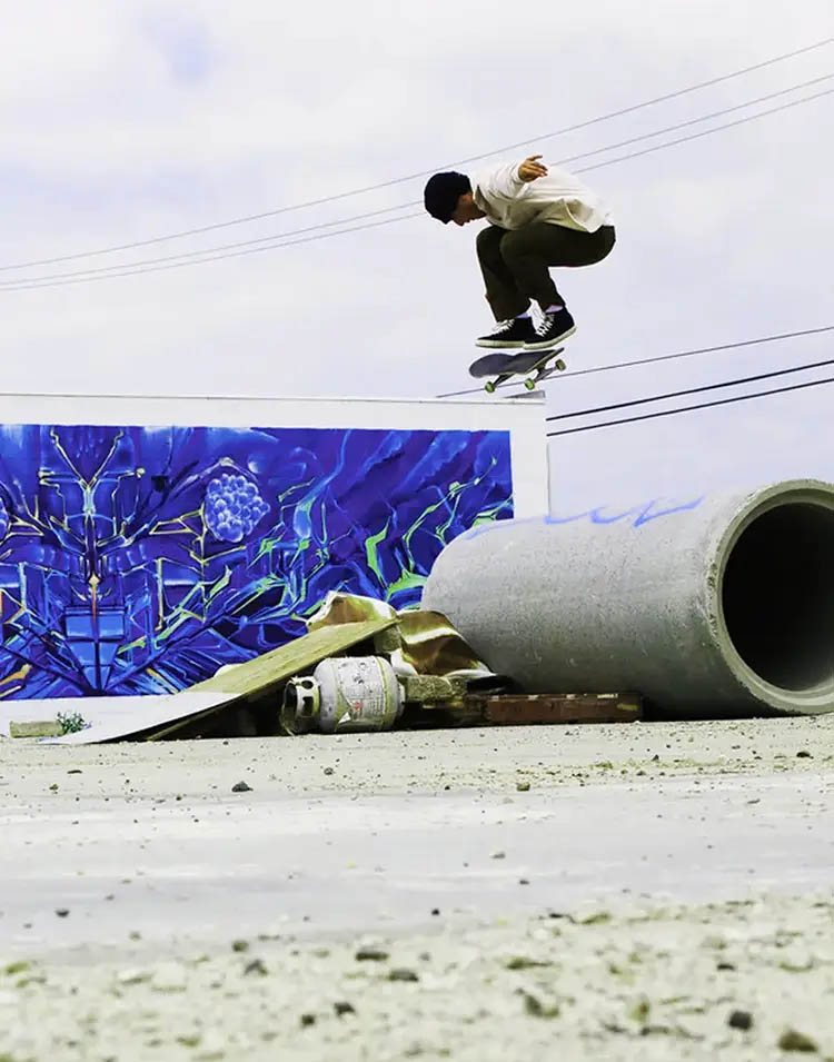 Photo of a skateboarder performing a Backside Kickflip over a concrete tube in front of a blue wall