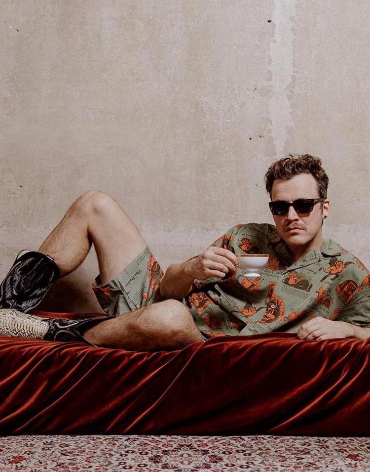 Picture showing a young white male laying on a red velvet daybed, holding a white porcelain cup and being dressed in a green allover printed short sleeve shirt with red illustrations and the matching shorts that says "The Dudes" on it. He is facing the camera, wearing sunglasses and some snake skin leather boots.