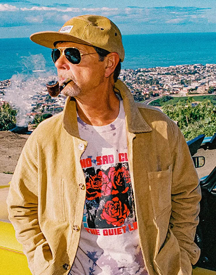Picture of a middle-aged man, smoking a pipe in front of a beautiful beach scene and wearing sunglasses, as well as a brownish cord outfit by The Quiet Life