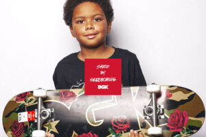 Portrait of a black child with a cheeky and proud smile, facing the camera and holding up a completely mounted skateboard by DGK with a camo inspired artwork showing roses and a big DGK logo. The center shows a red square with the words »Saved by Skateboarding«.