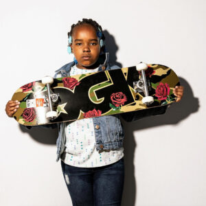 Portrait of a child facing the camera and holding up a completely mounted skateboard by DGK with a camo inspired artwork showing roses and a big DGK logo.