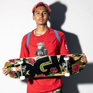 Portrait of a teenager facing the camera and holding up a completely mounted skateboard by DGK with a camo inspired artwork showing roses and a big DGK logo.