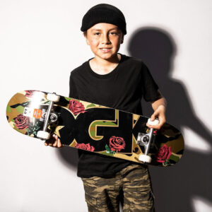 Portrait of a child facing the camera and holding up a completely mounted skateboard by DGK with a camo inspired artwork showing roses and a big DGK logo.