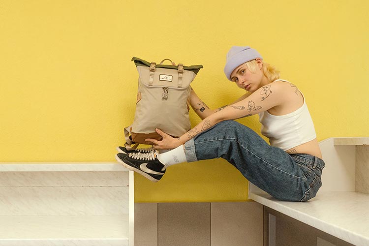 Portrait of a young women wearing a white top, purple beanie and blue jeans. She is facing the camera, sitting in front of a yellow wall, holding up a Doughnut backpack in front of her.