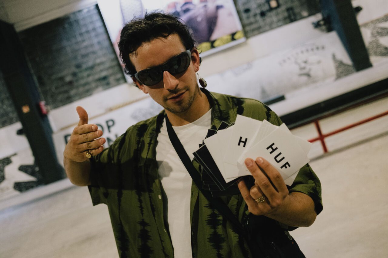 Photo showing the event’s host Rubens, wearing sunglasses and holding a bunch of white HUF stickers into the camera.