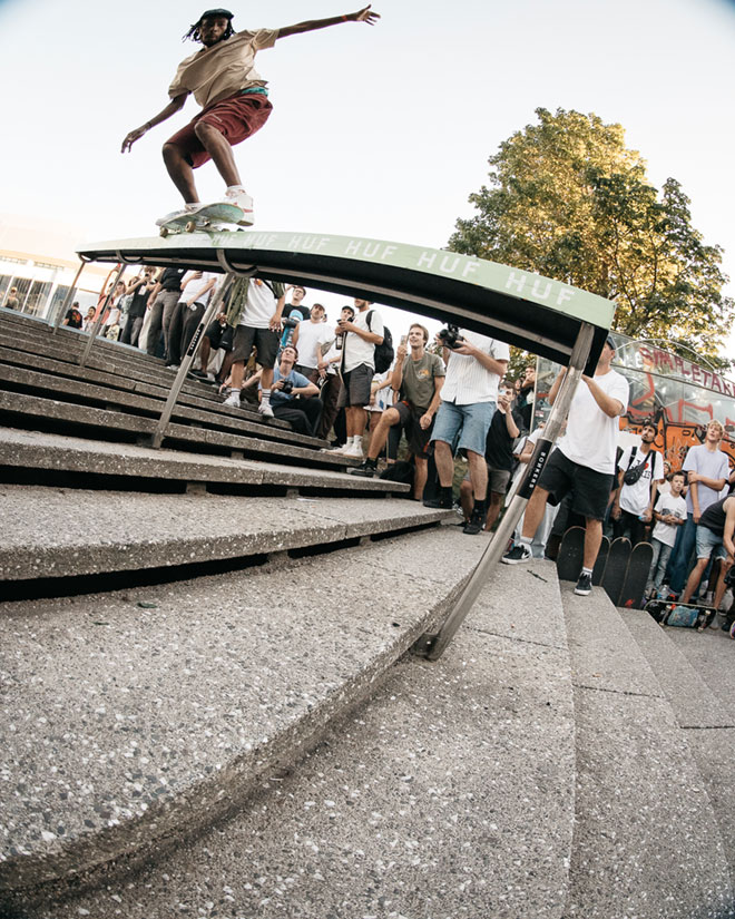 Photo of Kanya Spani doing a 50-50 grind down a gnarly handrail with a green topper during the HUF “Rail around the corner” event at SKTWK 2023.