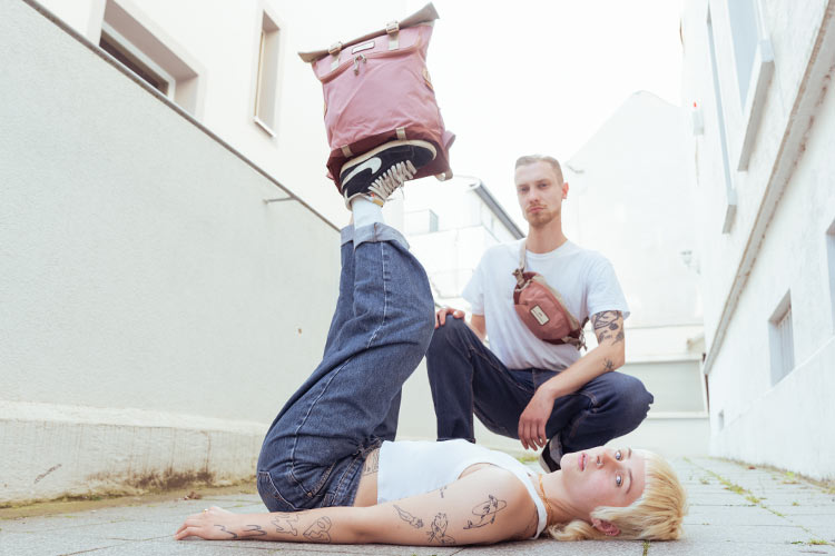 Young couple posing in front of a whiteish background. A short-haired blonde woman is laying on the ground, lifting her legs up and balancing a pastel pink backpack by Doughnut. A young male wearing a crossbody bag in the same color is squatting in the background, both facing the camera.