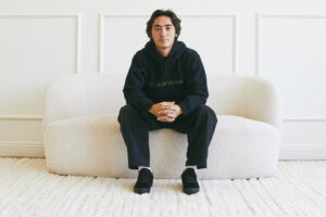 Photo of young professional skateboarder Gage Boyle, dressed in all black and sitting on a white vintage couch, looking straight into the camera.