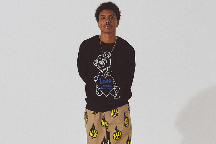 Young adult wearing a black HUF sweatshirt with a teddy bear print and the word "Love" printed on, facing the camera and having his arms behind his back.