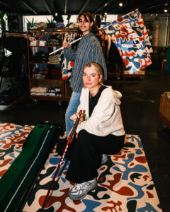 Portrait of two young women posing in front of a mini golf green and smiling into the camera. One is holding a golf putter and the other a flag with a colorful print and the PARLEZ logo.